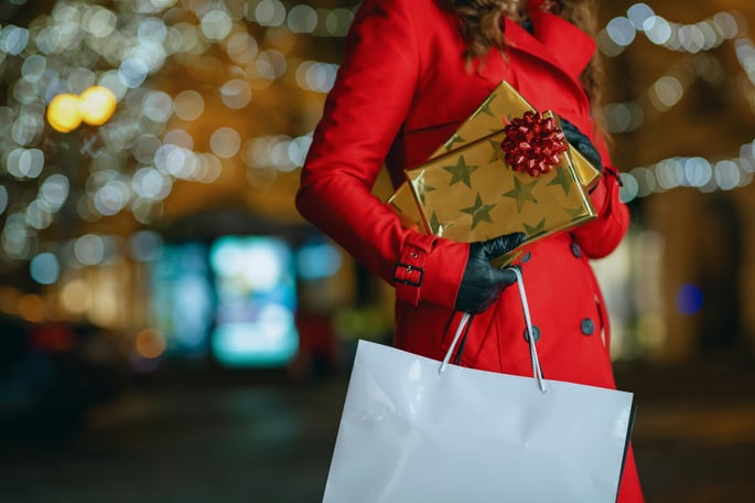 Woman in a red trench coat holding a white shopping bag and two wrapped presents.  