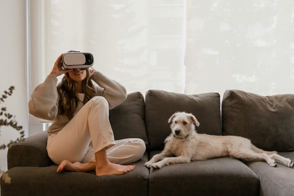 Woman sitting on the couch wearing a VR headset and her dog is sitting next to her.