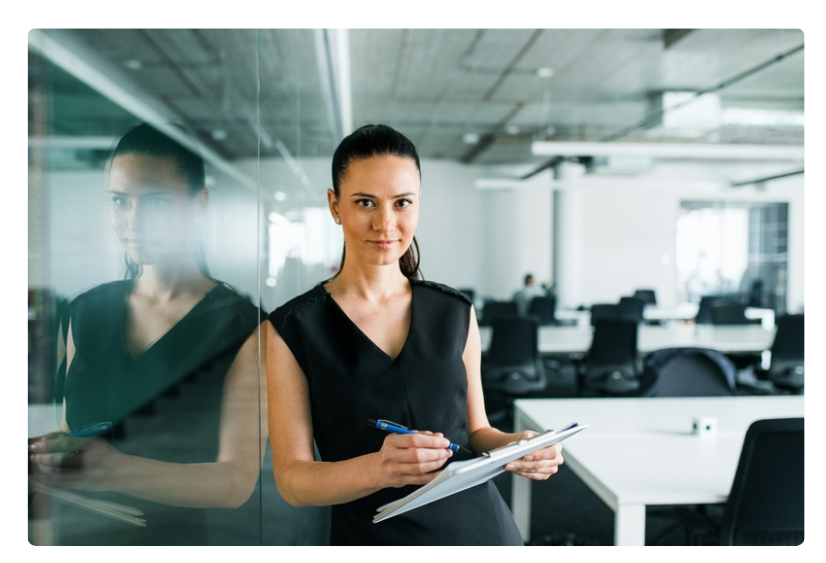 Woman standing in an office with a pad of paper and pen in her hand. She is wearing a black sleeveless dress. Woman standing in an office with a pad of paper and pen in her hand. She is wearing a black sleeveless dress.